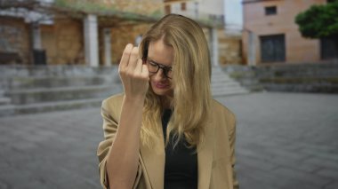 Açık havada, gözlüklü, sarı saçlı, spor ceketli, güneşli şehir ortamında gösteriş yapan bir kadın..