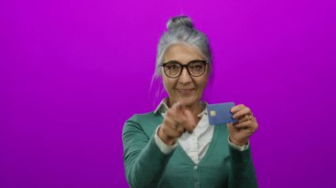 Gri saçlı, gülümseyen ve işaret eden yaşlı bir kadın kredi kartını soyutlanmış pembe arka planda tutuyor..