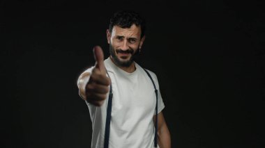 Pantolon askısı ve beyaz gömlek giyen İspanyol bir adam siyah arka plana karşı olumlu ve kendine güvenen neşeli bir gülümseme ve neşeli bir jestle başparmak kaldırıyor..