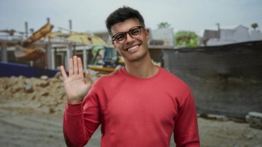 Şehirdeki inşaat alanında kırmızı tişörtlü bir genç açık havada dostça bir jest sergiliyor. Arka planda çekici bir portre sergiliyor..