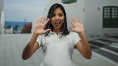 Güneşli caddede parlak mavi okyanus manzarası, neşeli atmosferi ve günlük kıyafetleriyle Latin cazibesini ve açık hava senaryosunu vurgulayan bir kadın..