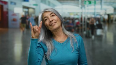 Gri saçlı, son sınıf öğrencisi bir kadın hareketli bir havaalanı terminalinde taş işareti yapıyor. Enerji ve seyahat ruhunu gösteriyor..