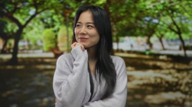 Yeşil bir parkta düşünceli bir şekilde gülümseyen genç bir kadın. Etrafı yemyeşil manzarayla çevrili..