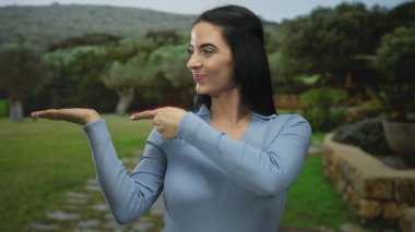 Parktaki genç İspanyol kadın başparmağıyla hareket ediyor ve gülümsüyor yemyeşil bir arka planda pozitif bir hava yaratıyor.