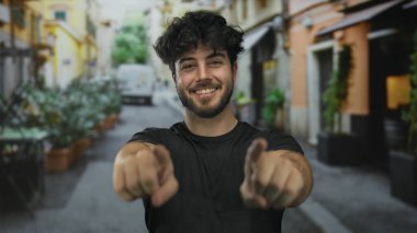 Sakallı genç adam gülümsüyor ve canlı bir sokakta dışarıya işaret ediyor..
