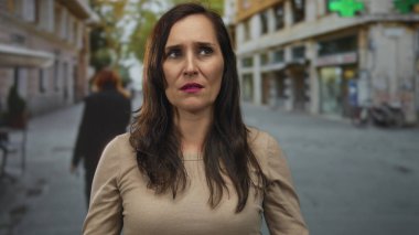 Şehir sokağında duran, arka planda binalar olan, bej bluz giyen, açık havada duygularını gösteren kafası karışık bir kadın..