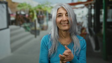 Gri saçlı bir kadın canlı bir sokakta el çırpıyor, mutluluk ve pozitifliği ifade ediyor, mavi süveter giyiyor, dükkanların ve kafelerin bulanık arka planında..