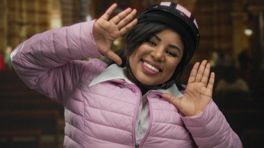 Kapalı alanda gülümseyen, kasklı pembe ceketli ve elleri havada bir kilisenin içinde sıcaklığı ve mutluluğu yansıtan bir jest yapan bir kadın..