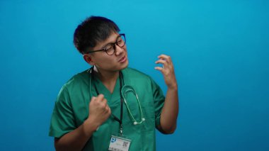 Yeşil önlüklü genç Çinli adam canlı mavi bir duvara karşı hava gitarı çalarken neşeli bir şekilde taklit ediyor, gözlük takıyor ve stetoskop takıyor, neşeli bir enerji yayıyor..