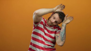 Çizgili tişörtlü genç İspanyol, izole edilmiş turuncu arka plana karşı neşeli kulak hareketi yapıyor..