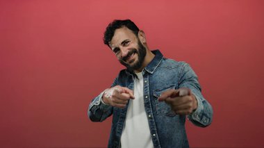 Sakallı İspanyol bir adam gülümsüyor ve kırmızı arka planda kot ceket giyiyor..