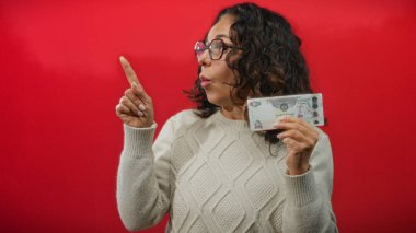 Orta yaşlı bir kadın elinde Uae Dirham banknotu, şaşırmış yüz ifadesi, kırmızı arka plan, gözlük ve süveter giyiyor,.