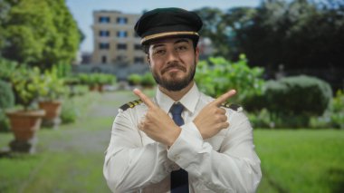 Pilot üniforması ve şapkası içinde sakallı, gülümseyen genç İspanyol adam yeşil bir parkta duruyor. Arka planda bulanık bir bina var..