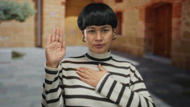 Açık havada duran kısa saçlı kadın şehir ortamında yemin ederken çizgili kazak ve ciddi bir ifade takınıyor..