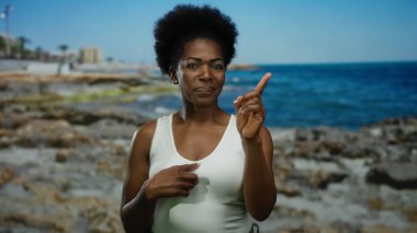 Mavi gökyüzünün altındaki kayalık plajda el kol hareketi yapan bir kadın, doğal deniz manzarası sergiliyor, ifadesini ve sahil ortamında dış görünüşünü vurguluyor..