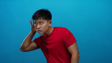 Kırmızı gömlekli genç adam stüdyo ortamında mavi arka plan duvarına karşı dikkatle dinliyor..