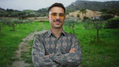 Genç, Latin asıllı bir adam, gür bir park manzarasında güvenle duruyor, kollarını kavuşturup gülümsüyor, şık gözlükler takıyor ve rahat bir açık hava havası yayıyor..