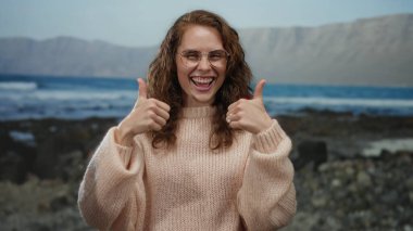 Deniz kenarında gülümseyen ve baş parmağını kaldıran bir kadın, gözlük takan, dağlık bir arka planda neşeli açık hava havasını vurgulayan bir kadın..