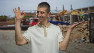 İnşaat alanındaki beyaz bir adam, beyaz bir polo giyip, inşaat malzemeleri ve makineleri arasında güvenle gülümseyen, memnuniyetini ve onayını ifade eden, beyaz bir jest yapıyor..