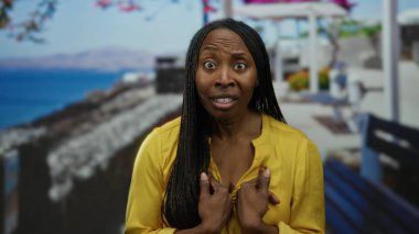 Sarı tişörtlü, uzun saçlı bir kadın güneşli bir günde deniz manzaralı bir sahilde dikiliyor..