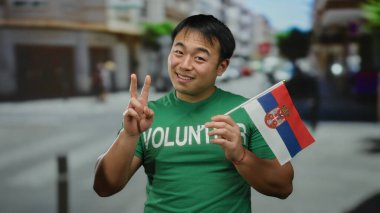 Kentsel caddede gönüllü tişört giyen genç adam barış işareti gösteriyor ve gülümsüyor, kentteki kültürel çeşitliliği simgeliyor..
