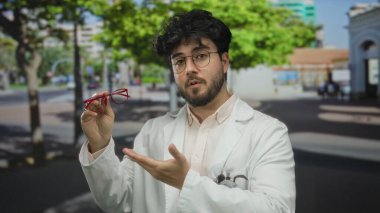 Beyaz önlüklü, sakallı, gözlüklü, kırmızı gözlüklü, güneşli bir sokakta arka planda ağaçlar ve binalar olan genç bir adam..