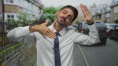 Beyaz tişörtlü ve donanma kravatlı bir adam elleri ile çerçevelenmiş, güneşli bir caddede dostça sırıtıyor ve kendinden emin bir duruş sergiliyor. Mutluluk..