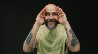 İzole edilmiş siyah bir arkaplan, sakallı, kel kafalı ve nötr renkli tişörtlü bir adama karşı şaşırmış pozlar veren adam belirgin özellikleri vurguluyor..