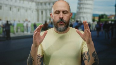 Dışarıdaki pisa kulesinin önünde sakince meditasyon yapan adam sakin bir sokak ortamında dövmelerini ve sakallarını sergiliyor..