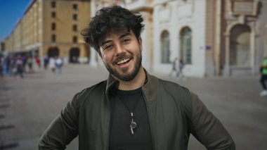 Yoğun bir caddede sakallı genç bir adam güvenli bir şekilde gülümsüyor tarihi binalar ve arka planda yürüyen insanlar kentsel yaşam tarzını ve kültürünü gözler önüne seriyor..