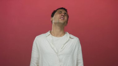 Beyaz gömlekli genç adam kırmızı arka planda duruyor. Duygusal ifadesini kapalı gözlerle gösteriyor.