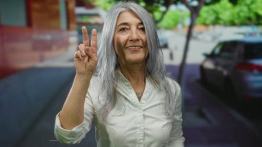 Beyaz gömlekli, kır saçlı yaşlı bir kadın sokakta dikiliyor. Ağaçların ve arabaların kentsel arka planında barış işareti var..