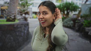 Yeşil bir çevre ile çevrili, bir park ortamında gülümseyen kadın, neşeli bir ifade ve el kulağına jest ile günlük kıyafet giyerek.