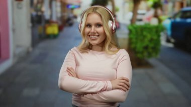 Kulaklık takan sarışın bir kadın canlı bir şehir caddesinde kollarını kavuşturmuş, açık havada genç enerjisini ve tarzını somutlaştırıyor..