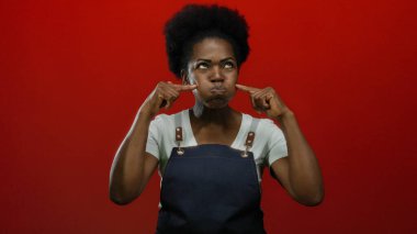 Önlüklü Afrikalı Amerikalı bir kadın parmaklarını parlak, izole kırmızı bir arka plana koyarken şakacı bir şekilde yanaklarını şişiriyor..