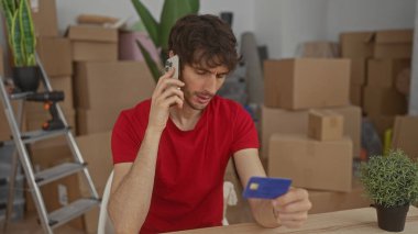 Adam telefonu kulağına tutuyor ve binanın içindeki kutular arasında kredi kartını elinde tutuyor..