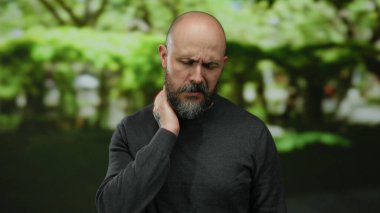 Dövmeli sakallı kel adam güneşli bir günde yemyeşil bir parkta boğazını tutmaktan rahatsız görünüyor..