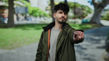 Sakallı genç adam, arka planda kararlılık ve odaklanma gösteren ağaçlar ve binalarla dolu bir parkta parkı gösteren bir ceket giyiyor..