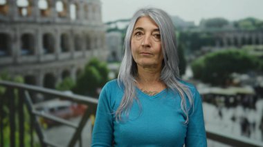 Mavi kazaklı, gri saçlı kadın Roma, İtalya 'daki ikonik Roma kolezyumunun yanında dikiliyor. Etrafı tarihi mimari ve yeşilliklerle çevrili..
