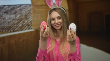 Tavşan kulaklı genç bir kadın, tarihi bir üniversite binasında renkli paskalya yumurtaları tutuyor. Kapalı alanda neşeli, neşeli bir atmosfer sergiliyor..