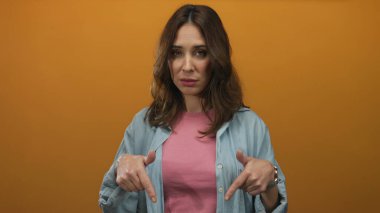 İzole edilmiş turuncu arka plana bakan bir kadın genç İspanyol kadını işaret ediyor. Ciddi ifade. Sıradan giyinen kot ceket kolları el ele..