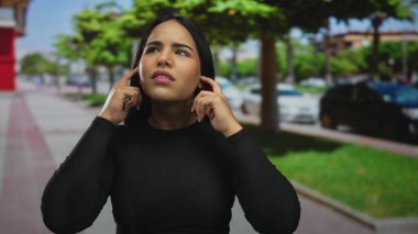 Kadın, kalabalık bir sokakta kulaklarını tutuyor ve Latin kökenli genç adam gürültüden rahatsız oluyor..