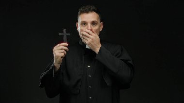 Rahip gibi giyinmiş bir adam karanlık bir arka plana karşı elinde haç, ağzının üzerinde bir el, sürpriz ya da şok, izole edilmiş ve gizemli dini imgeler iletiyor..