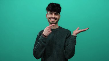 Genç bir adam yeşil bir arka planda gülümseyerek avucunun içini işaret ediyor, kendine güveni ve cazibesi ile süveter giymiş sıradan bir stile sahip..
