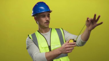 Yansıtıcı yelek giyen genç İspanyol bir adam ve izole edilmiş sarı arka plan duvarına bantlı kasklı ölçüler inşaat işi öneriyor..