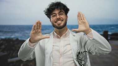 Sakallı ve gözlüklü genç adam güneşli bir günde arka planda deniz kenarında el kol hareketleri yapıyor.