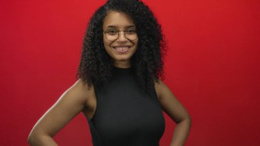 Kıvırcık saçlı ve gözlüklü bir kadın canlı kırmızı bir arka plan önünde gülümsüyor, kendine güveni ve sıcaklığı genç ve dinamik bir ortamda somutlaştırıyor..
