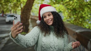 Noel Baba şapkası takan bir kadın güneşli bir caddede açık havada selfie çekiyor ağaçlar ve arabalarla çevrili, gezici cihazıyla neşeli tatil anlarını yakalıyor..