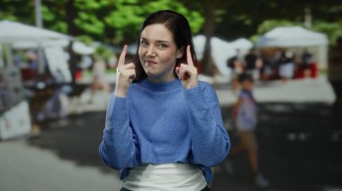 Güneşli bir günde, açık havada mavi kazaklı bir kadın arka planda market tezgahlarıyla yukarı bakıyor. Merakını dile getiriyor ve hareketli bir sokak sahnesine odaklanıyor..