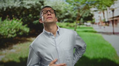 Yüzünde acılı bir ifadeyle güneşli yeşil bir parkta dikilirken midesi ağrıyan gözlüklü beyaz bir adam..